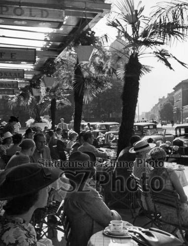 Ein überfülltes Straßencafé mit Menschen, die an einem sonnigen Tag unter Palmen sitzen, Oldtimer auf der Straße.