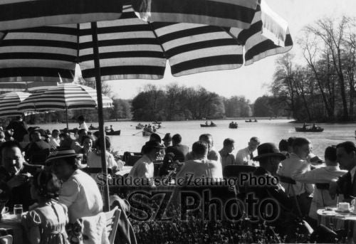 Volles Straßencafé an einem See mit gestreiften Sonnenschirmen, Menschen, die essen, und Booten auf dem Wasser im Hintergrund.