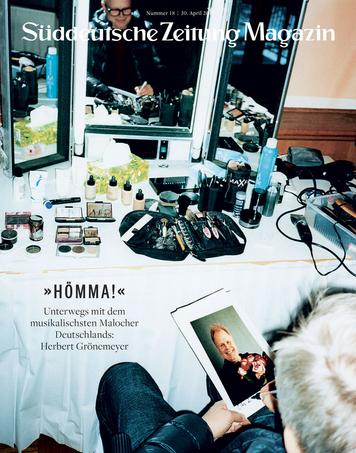 Ein überladener Schminktisch mit Kosmetika; eine Person hält ein gerahmtes Foto und schaut in einen Spiegel.