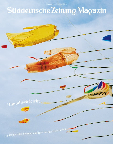 Bunte Stoffdrachen in Form von Kleidern fliegen vor einem blauen Himmel mit Wolken und fließenden Bändern.