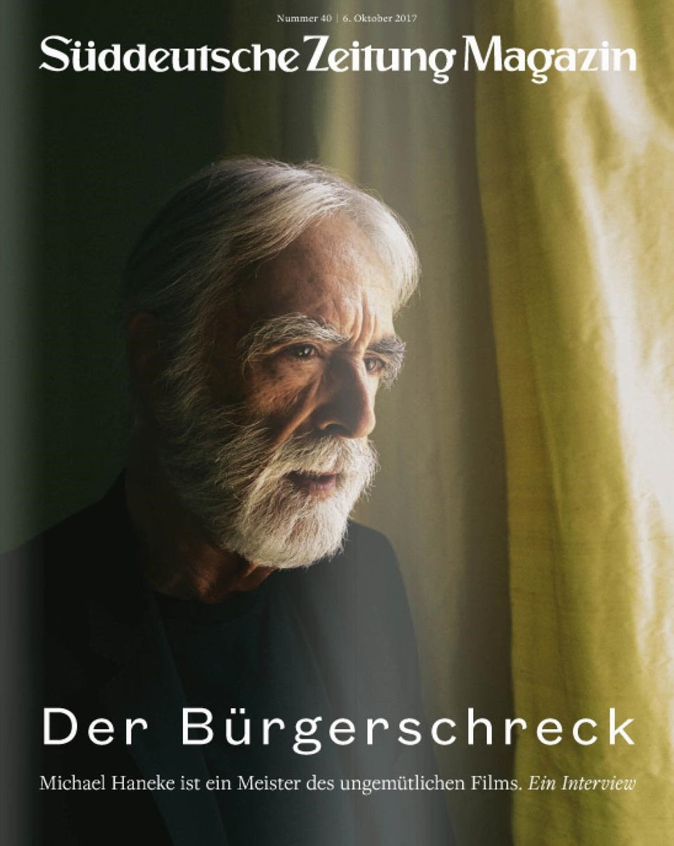 Ein älterer Mann mit weißem Bart steht an einem Fenster mit gelben Vorhängen und blickt ins Licht.
