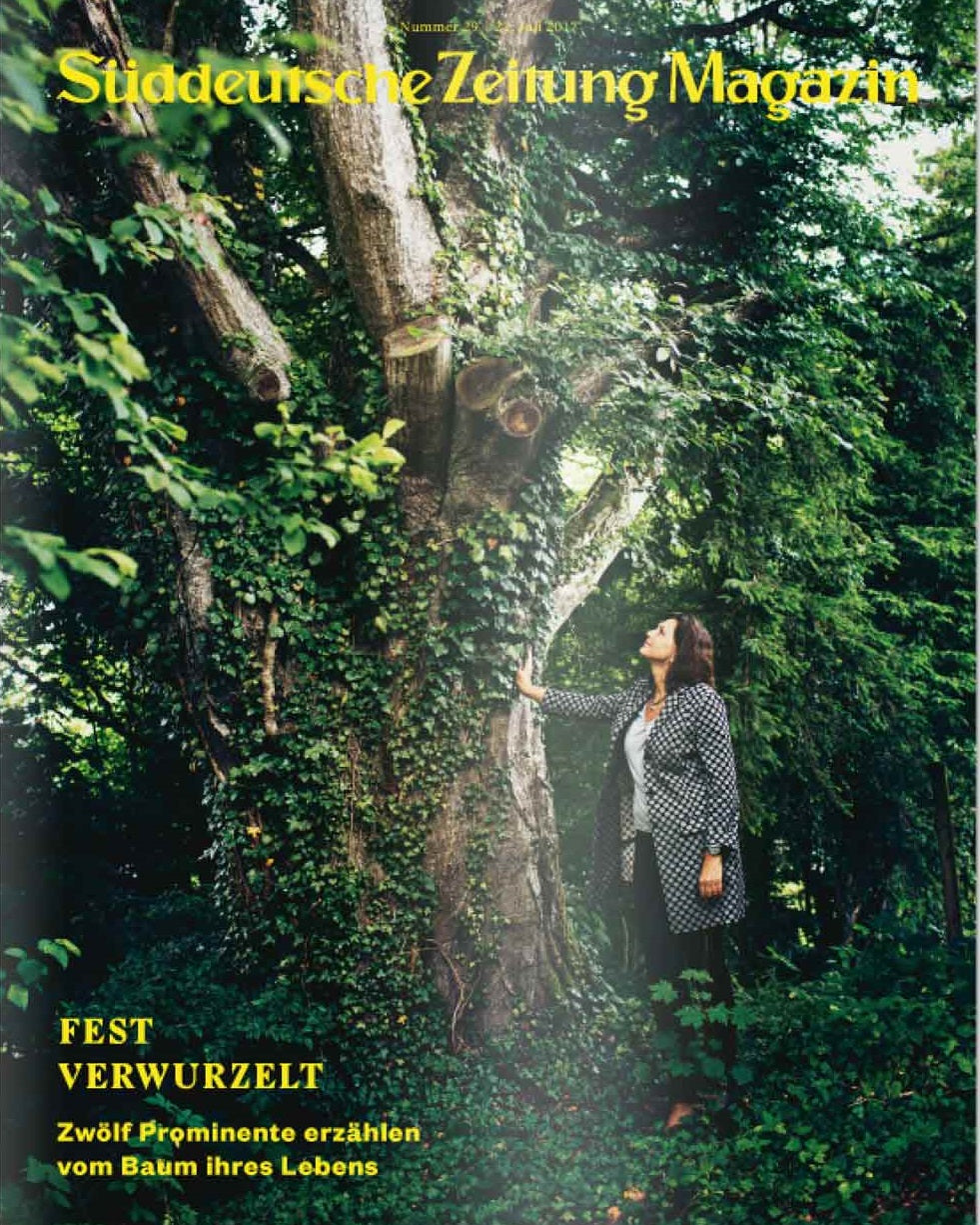 Auf dem Titelbild einer Zeitschrift steht eine Frau, die einen großen Baum berührt, umgeben von üppigem Grün.