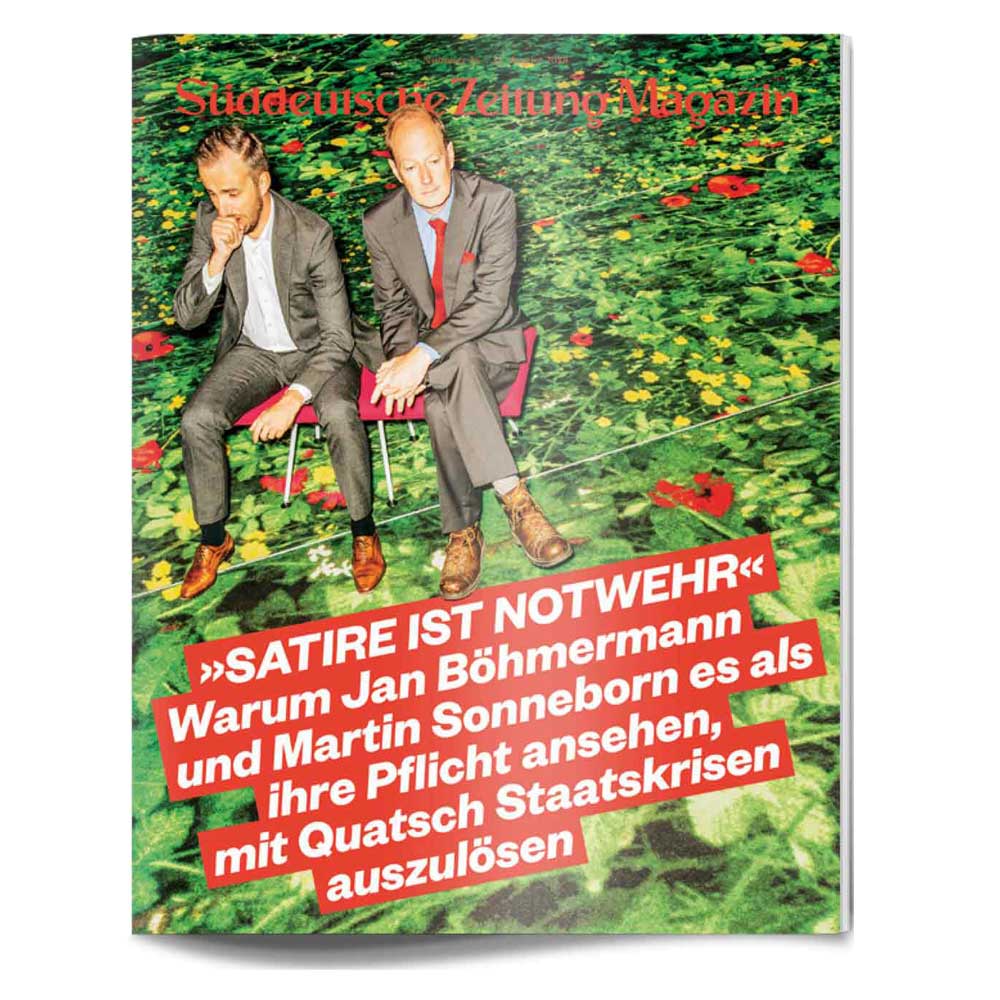 Zwei Männer in Anzügen sitzen auf roten Stühlen auf einer Wiese mit Blumen, Süddeutsche Zeitung Magazin Titeltext in Deutsch.